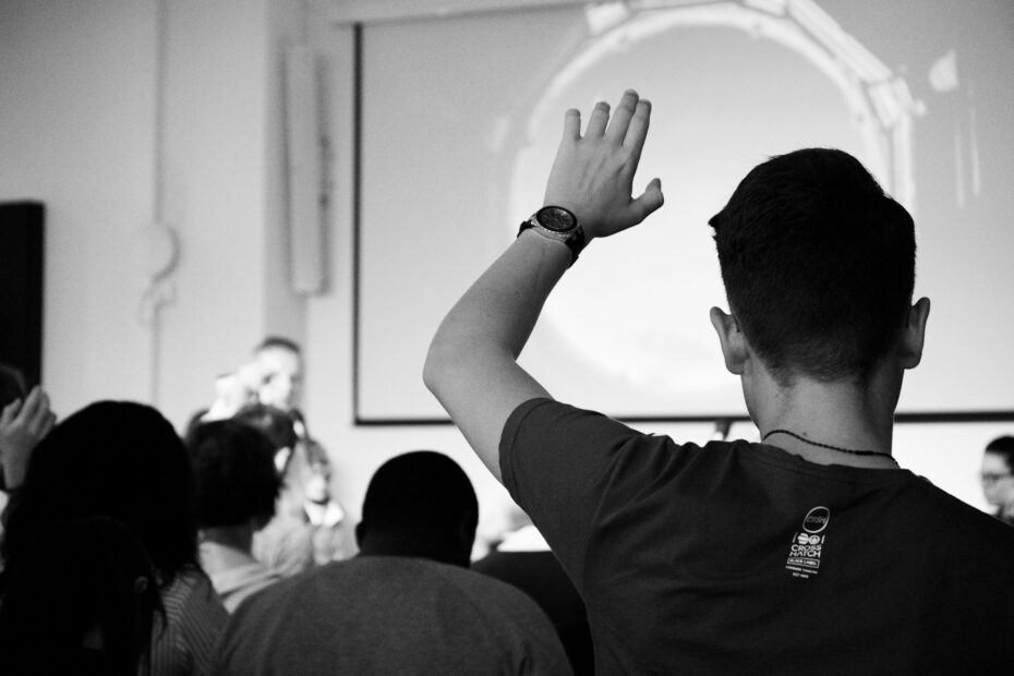 An image in black and white for a man holding his hand to ask a question in some lecture.