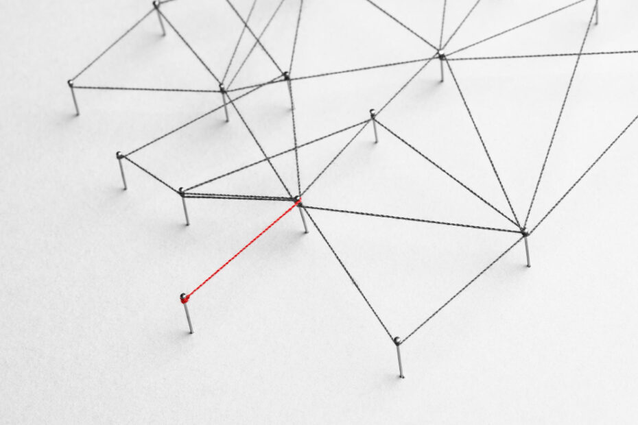 A network representation using black strings connected by small metal pins on a white surface, forming a complex web-like structure. One string is highlighted in red, emphasizing a specific connection within the network. This abstract visualization depicts interconnected nodes, often used to illustrate concepts like data structures, networks, or relationships in technology and science.