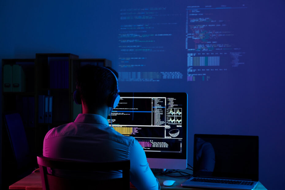 A software developer is sitting at a desk in a dimly lit room, working on code displayed on a large monitor. The screen shows multiple lines of code and data visualizations, indicating a focus on programming or software development. The developer is wearing headphones, suggesting deep concentration or possibly listening to music while working. Additional code is projected onto the wall in the background, creating a tech-focused, immersive atmosphere. The setup includes a laptop beside the desktop monitor, reinforcing the multitasking nature of the work environment.