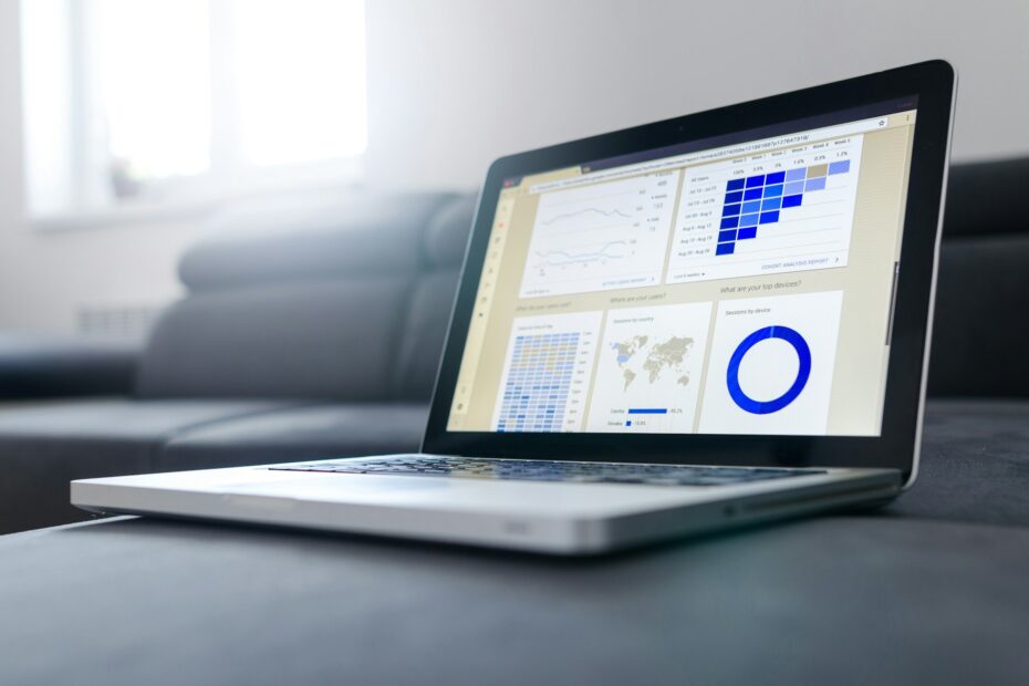 A laptop displaying various analytical charts and graphs on its screen, including bar graphs, pie charts, and line graphs, all in blue tones. The laptop is placed on a couch in a bright room, with natural light coming through a window in the background. The screen shows data analysis visuals, likely related to business or marketing metrics.