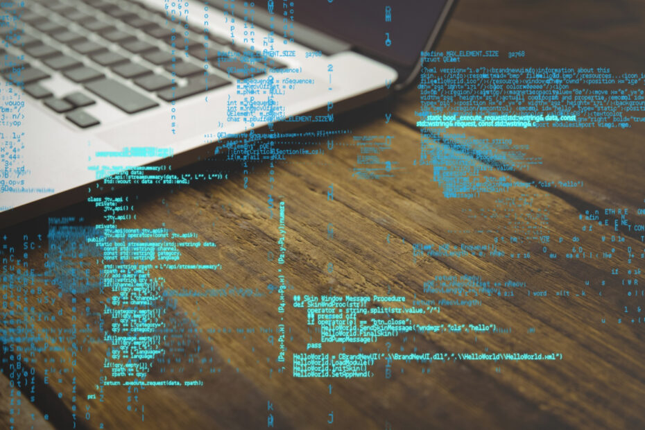 A close-up of a laptop on a wooden desk with blue programming code overlaid digitally, symbolizing web development. The image represents coding in JavaScript frameworks like React and Vue, commonly used for building modern web applications.